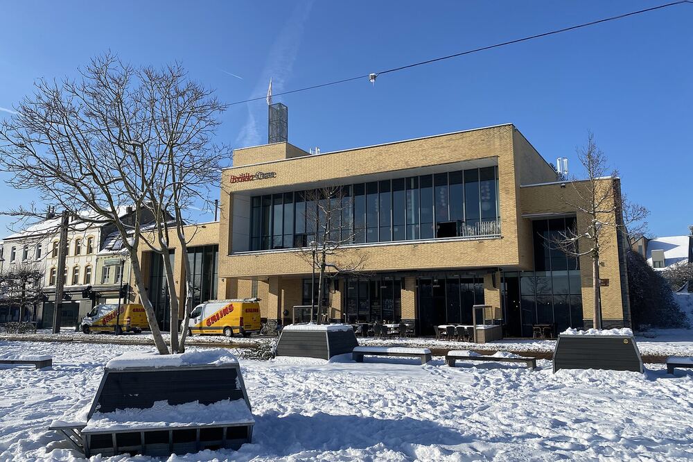 Het gele bakstenen gebouw van cultureel centrum Brikke Oave in Brunssum onder een strakblauwe winterhemel, met een besneeuwd plein met banken en kale bomen ervoor